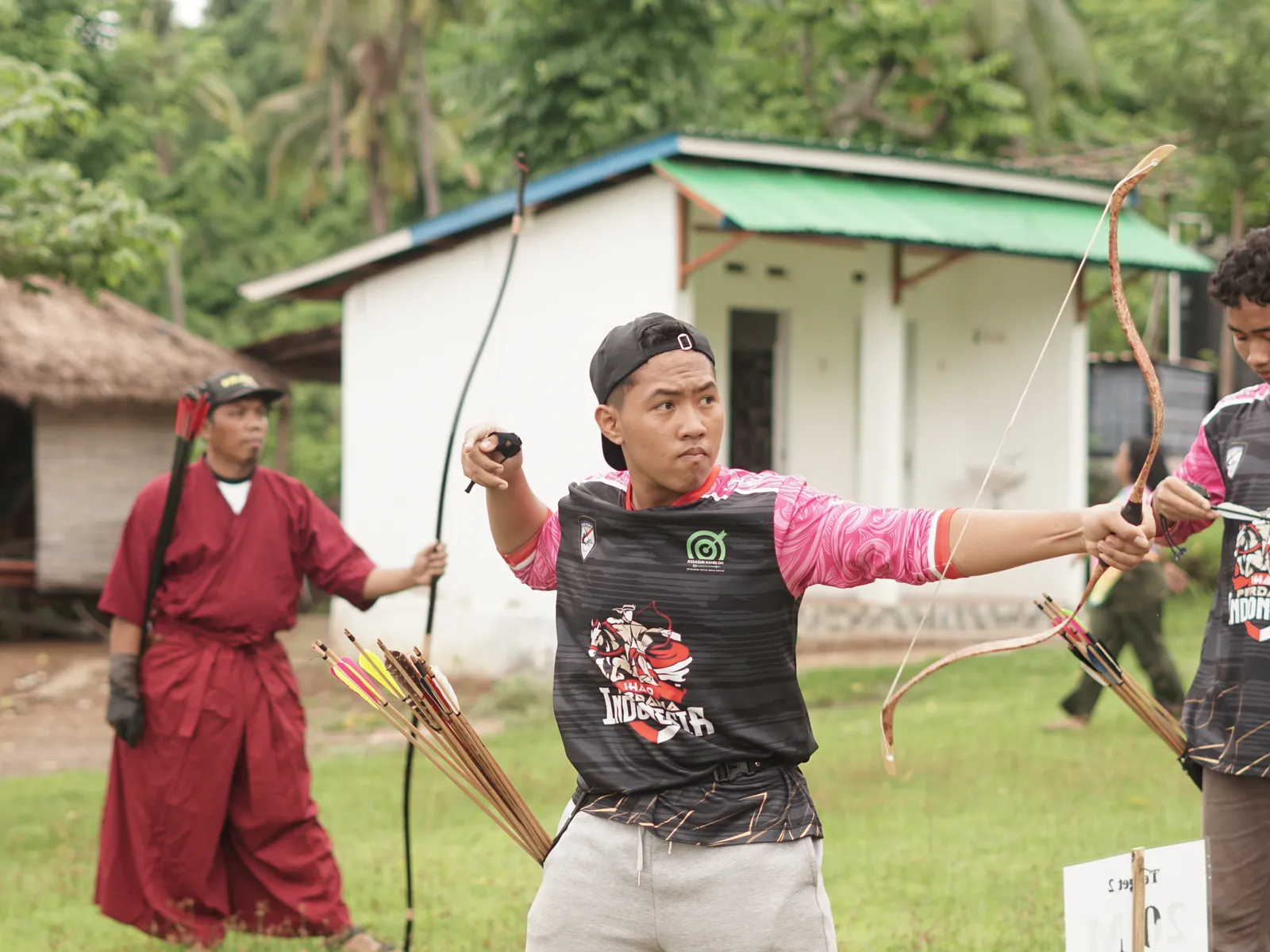 Archer in pink and black uniform drawing traditional wooden bow during IWFA competition with traditional building in background