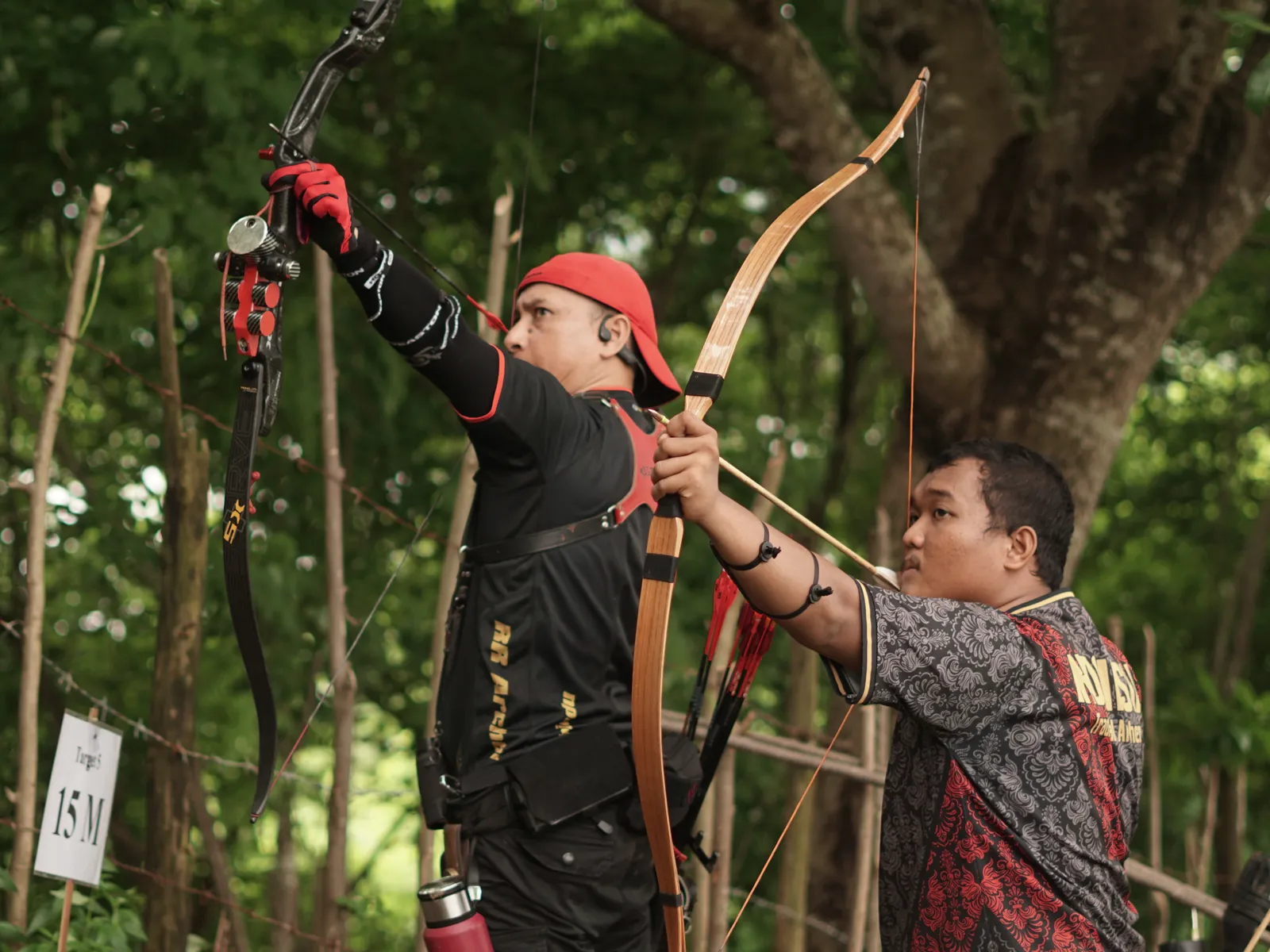Two archers competing, one in red headband with traditional wooden bow, another in patterned shirt in forest setting
